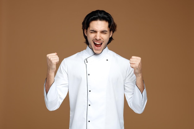 frowning young male chef wearing uniform showing yes gesture screaming with closed eyes isolated on brown background