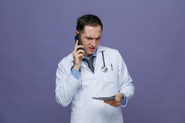 Хмурый молодой врач-мужчина в медицинском халате и стетоскопе на шее держит деньги, глядя на них, глядя вниз во время разговора по телефону на фиолетовом фоне