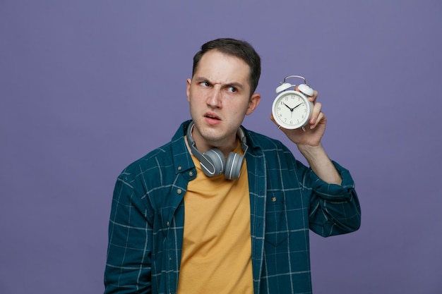 Frowning young male student wearing headphones around neck holding alarm clock near ear looking at side listening isolated on purple background