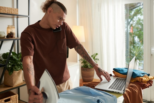 Frowning young man checking emails on laptop when ironing clothes at home