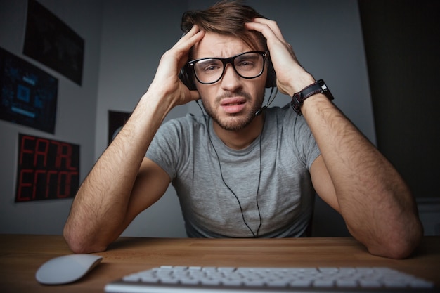 Foto giovane accigliato con gli auricolari e gli occhiali seduto con le mani sulla testa davanti al computer