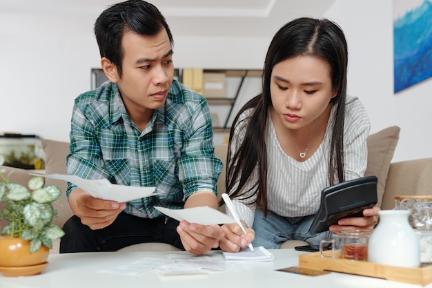 Frowning young man showing utility bill or shop receipt to his girlfriend who is managing finances