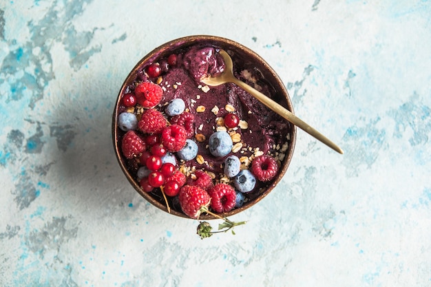 Frozen acai smoothie in coconut shell with raspberries, banana, blueberries,  fruit and granola on concrete background. Breakfast, healthy meal for summer vibes, top view, space for text