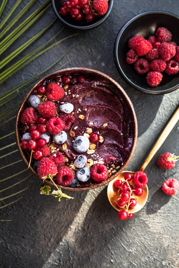 Frozen acai smoothie in coconut shell with raspberries, banana, blueberries,  fruit and granola on concrete background. Breakfast, healthy meal for summer vibes, top view, space for text