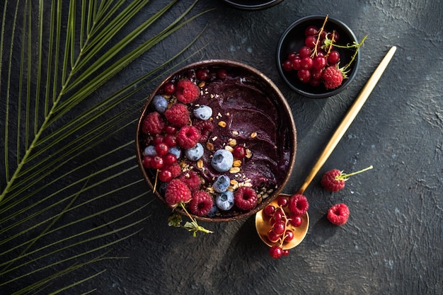 Frozen acai smoothie in coconut shell with raspberries, banana, blueberries,  fruit and granola on concrete background. Breakfast, healthy meal for summer vibes, top view, space for text