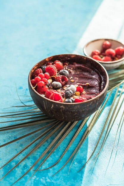 Frozen acai smoothie in coconut shell with raspberries, banana, blueberries,  fruit and granola on concrete background. Breakfast, healthy meal for summer vibes, top view, space for text