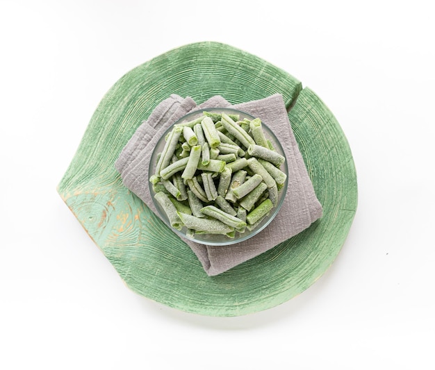 Frozen cut green beans in glass bowl on gray napkin on green wooden slice on white top view