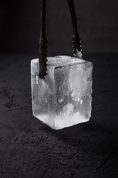 A frozen ice cube for rocks glass in bartender ice tongs, dark backdrop