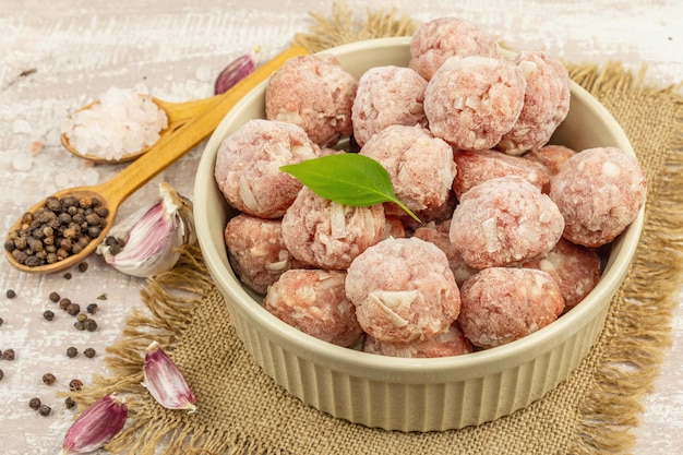 Frozen meatballs with spices, basil leaves, and garlic cloves in a ceramic bowl. Raw ingredients for cooking food. Wooden background, close up