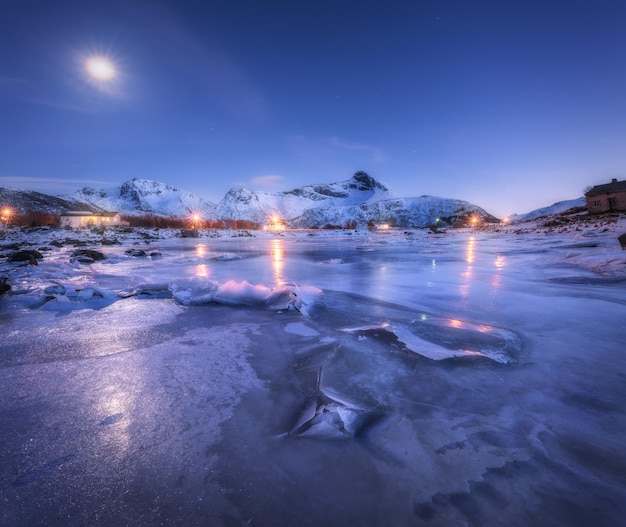 Frozen sea coast beautiful snow covered mountains and starry sky with moon in winter at night Beautiful fjord in Lofoten Islands Norway Nordic landscape with ice rocks buildings illumination