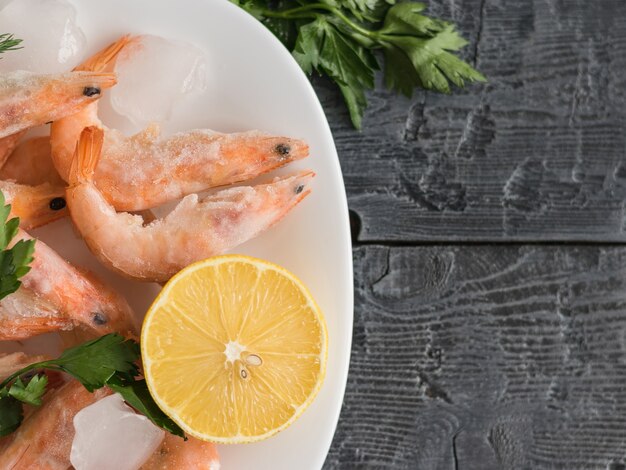 Frozen shrimp with lemon and herbs on white bowl on black wooden rustic table Top view