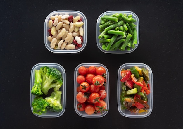 Frozen vegetables in plastic containers on black background. Frozen broccoli, green beans, cherry tomatoes, beans and bell pepper.