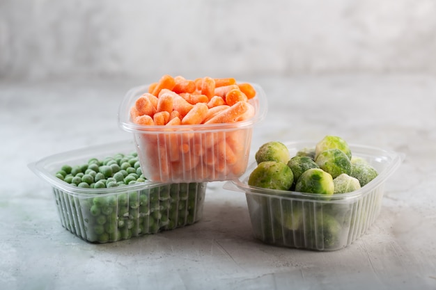 Замороженные овощи, такие как зеленый горошек, брюссельская капуста и морковь в пластиковых коробках на бетонном сером пространстве