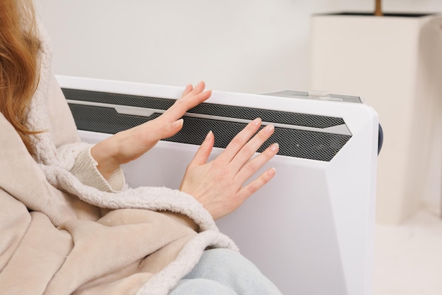 Frozen young woman in mittens warming near radiator at home closeup