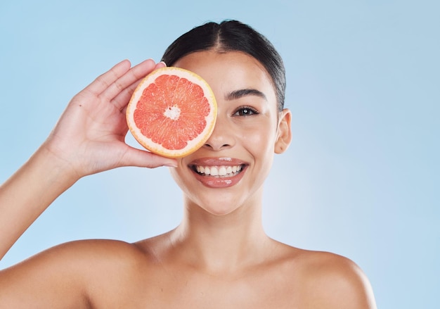 Bellezza della frutta e pelle di una donna con un sorriso che tiene un pompelmo sul viso su uno sfondo blu per studio ritratto di una modella felice in una cura della pelle sana, succosa e naturale per l'assistenza sanitaria