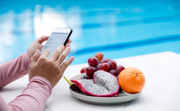 Fruit in dienblad met de hand van een Aziatische vrouw in een zwempak bij het hotelzwembad Glijdende mobiele telefoon