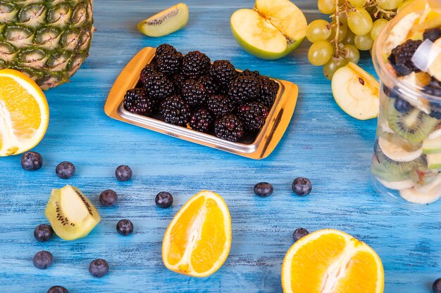 Fruit salad in plastic cup next to berried and other exotic fruits on wooden table. Delicious colorfull summer fuits dessert