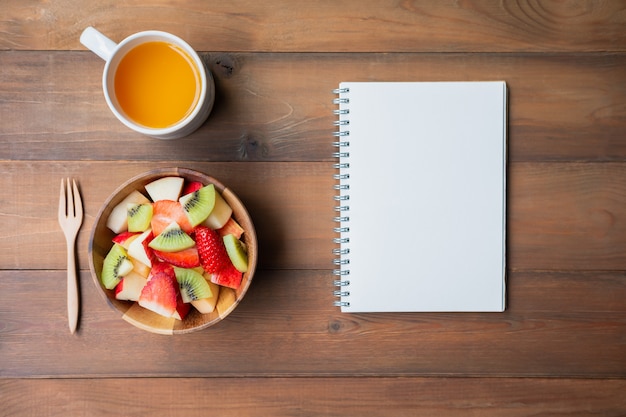 Fruit salad with orange juice and notebook on brown wood background with copy space,Flat lay.