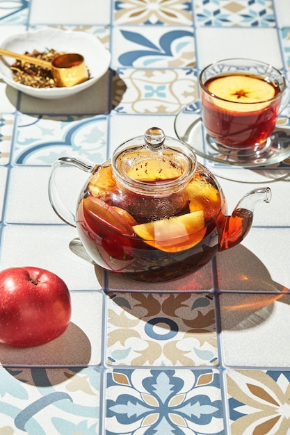 Fruit tea with apples and thyme in glass teapot and cup on table made of colored tiles