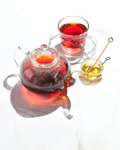 유리 찻주전자에 사과, 백리향, 꿀을 넣은 과일 차와 단단한 그림자가 있는 흰색 배경에 컵