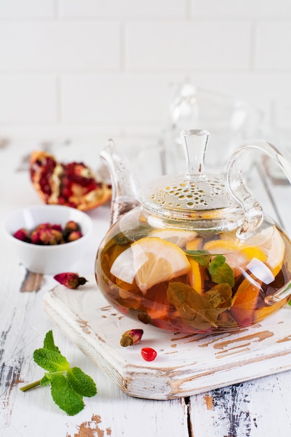 Fruit tea with berries, lemon, lime and mint leaves in glass teapot on white light wooden background. Selective focus.