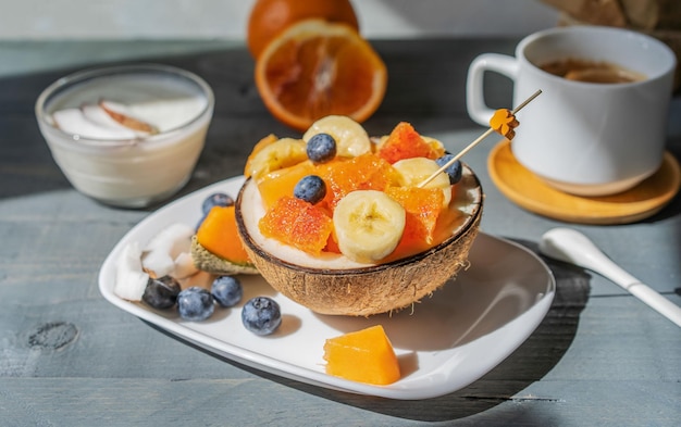 Fruit tropical salad in half of fresh coconut with cup of coffee on grey table