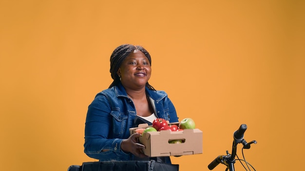 Fruitmand wordt voorzichtig uit de voedselbezorgzak gedragen door Afro-Amerikaanse fietskoerier. Vrouwelijke Afro-Amerikaanse koerier fietst terwijl ze grijnst terwijl ze verse producten levert.