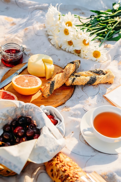 果物、クロワッサン、ジャム、紅茶、夏の日差しの中でテーブルクロスの上の花。ピクニックのコンセプト