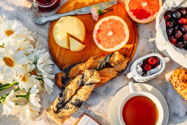 Fruits, croissants, tea and flowers on tablecloth in summer sunlight. Picnic concept