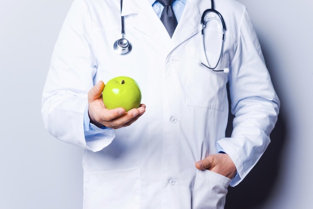 Fruits make you healthy. Close-up of doctor holding green apple while standing against grey background