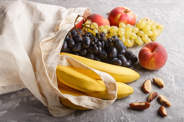 Fruits in reusable cotton textile white bag on a gray concrete background. Zero waste shopping, storage and recycling concept. Side view, close up