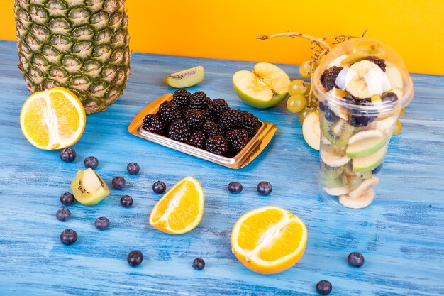 Fruitsalade in plastic beker naast bessen en ander exotisch fruit op houten tafel. Heerlijk kleurrijk zomers dessert