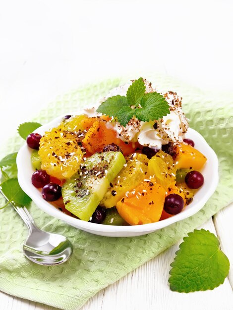 Fruitsalade met sinaasappel, kiwi, veenbessen en gebakken pompoen, slagroom, besprenkeld met chocolade en kokosnoot met munt in een kom op een servet op de achtergrond van licht houten bord