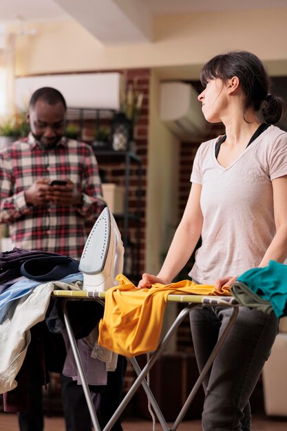 Frustrated angry housewife ironing clothes on board while african american husband not helping. Stressed tired unhappy modern woman doing everything without help.