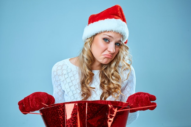 Frustrated and annoyed beautiful young woman in Santa Claus hat with a gift on blue