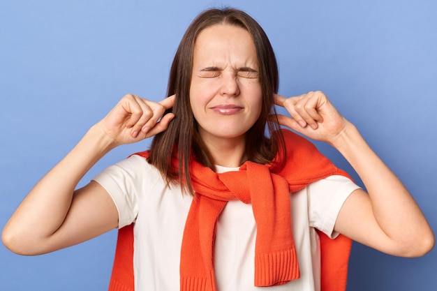 Frustrated annoyed woman in Tshirt and sweater tied over shoulders covering ears with hands feeling irritated with loud annoying noise can't concentrate on her work isolated on blue background