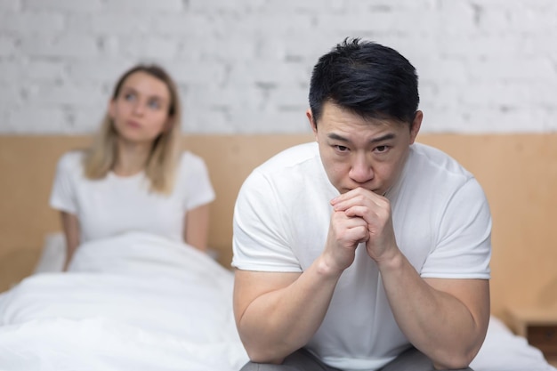 Разочарованная азиатка, сидящая на краю кровати, разочарованная семья и ссорящаяся в постели с несчастной семьей дома