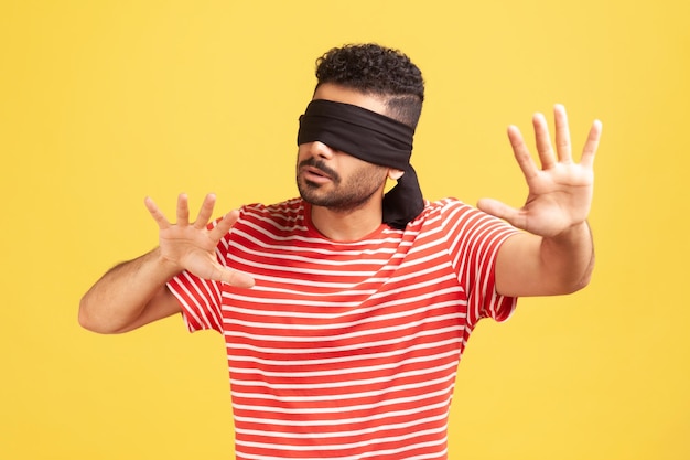 Frustrated bearded man in striped t-shirt with blindfold on his eyes trying to move in darkness touching space with hands, vision problems. Indoor studio shot isolated on yellow background