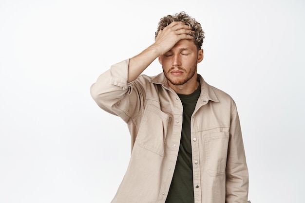 Frustrated blond man facing failure facepalm and look distressed being in trouble standing upset against white background
