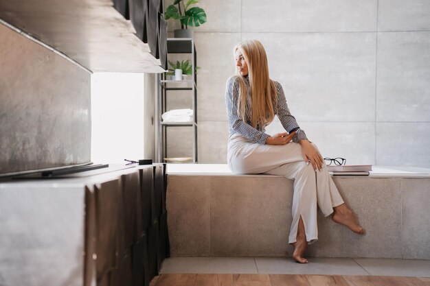 Frustrated blonde fit young woman in white pants and shirt holding phone sitting on bath at bathroom
