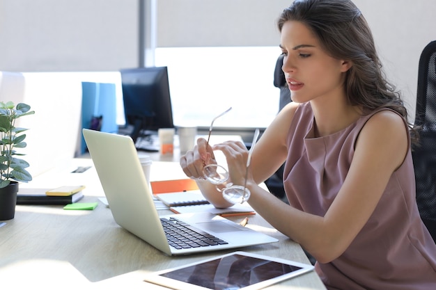 Frustrated business woman looking exhausted while sitting at her working place and carrying her glasses in hand.