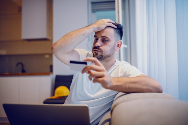 Frustrated caucasian handsome man in pajamas sitting in living room with laptop in lap and credit card in hand.