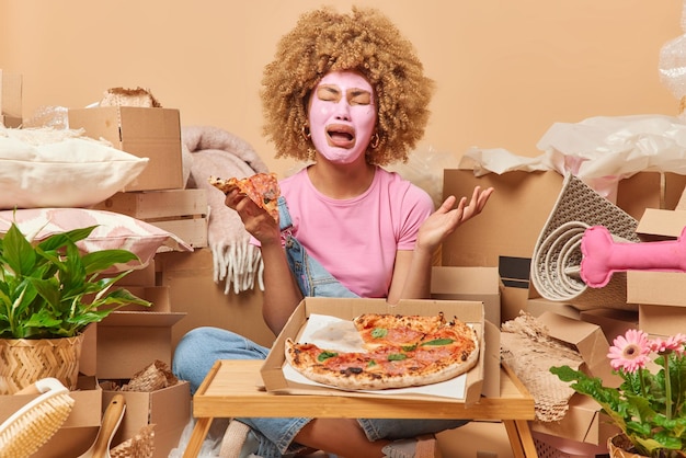 Frustrated crying woman applies beauty mask on face to reduce\
blackheads eats appetizing pizza sits crossed legs in empty room\
full of cardboard boxes full of household items relocation\
concept