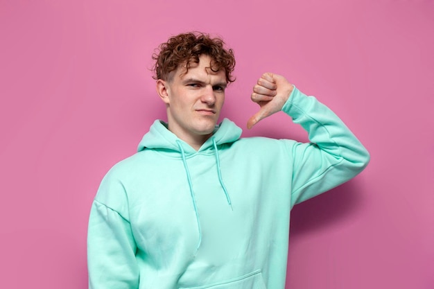 Frustrated curly guy in mint hoodie shows dislike on pink isolated background displeased man