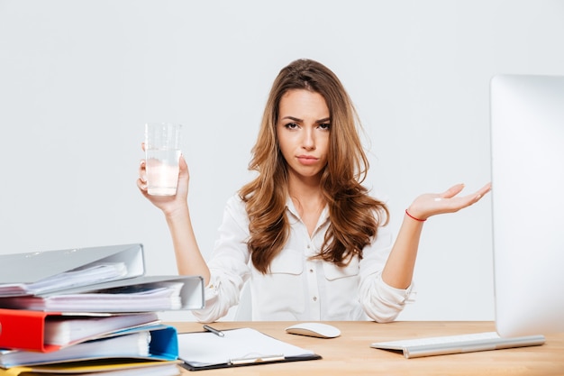 Разочарованная и разочарованная бизнесвумен, сидящая на дест в офисе с изолированным стаканом воды на белом фоне