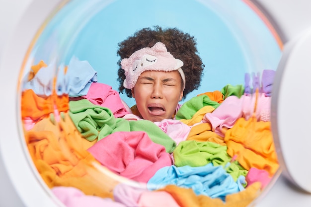 Frustrated disappointed curly haired tired woman cries from\
tiredness wears blindfold on forehead wants to sleep but has to\
finish housework poses in washing machine drum surrounded by\
laundry