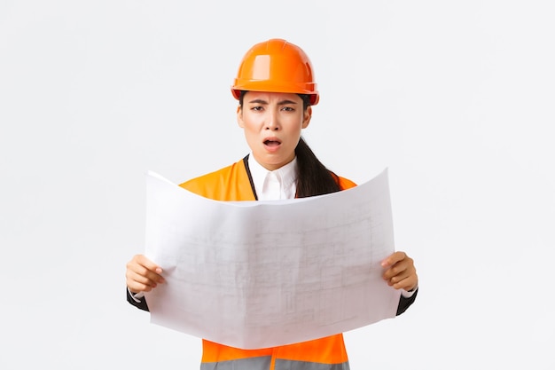 Frustrated and displeased female asian engineer, chief architect at construction zone, looking disappointed after studying blueprints, stare camera, scolding manager, white background