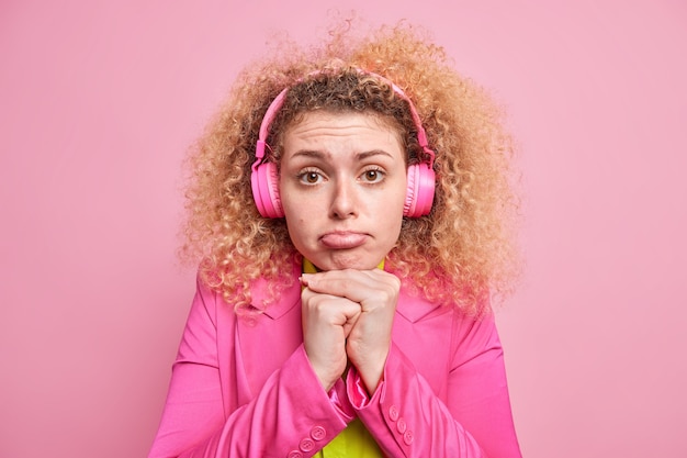 Frustrated European woman keeps hands under chin purses lips looks with sorrowful expression listens music via headphones dressed in bright clothes has bad mood isolated over pink wall