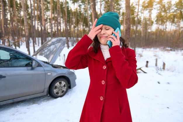 Frustrated female driver in tears of fright talking on mobile phone calling for roadside assistance standing on a snowy road in front of her auto with open hood Travel by car and breakdown concept