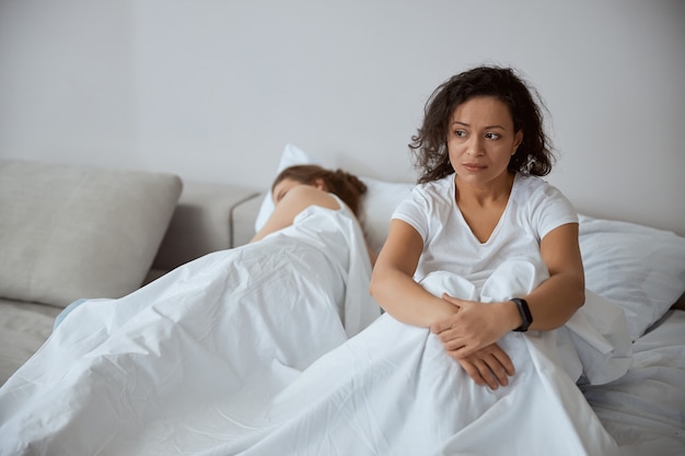 Frustrated female person sitting on the bed and embracing knees, looking aside